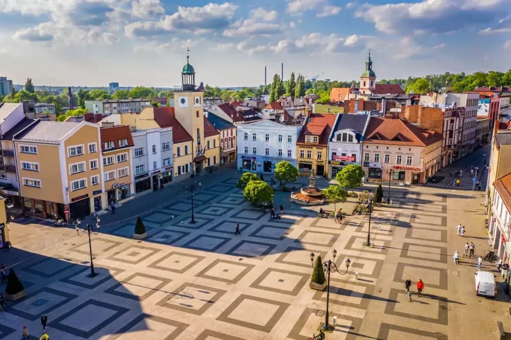 An aerial view of a town square with historic buildings, a prominent clock tower, and a decorative fountain, all under a blue sky with some clouds.